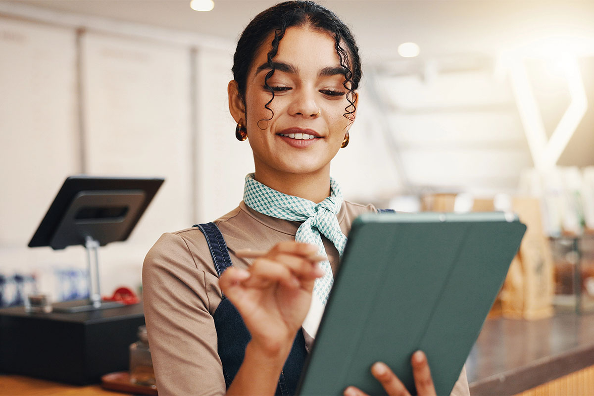 A young woman with curly hair and a scarf is smiling while using a tablet in a café setting.